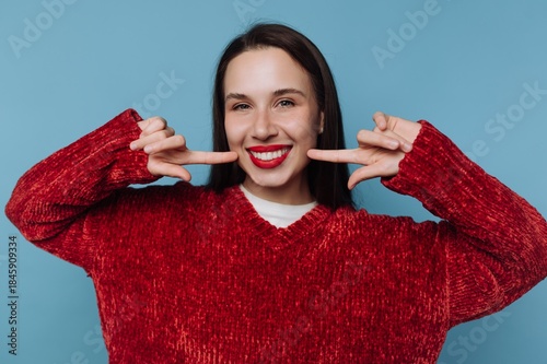 Woman smiles and poses with fingers on a blue background wearing a red sweater and bright lipstick while showing her teeth