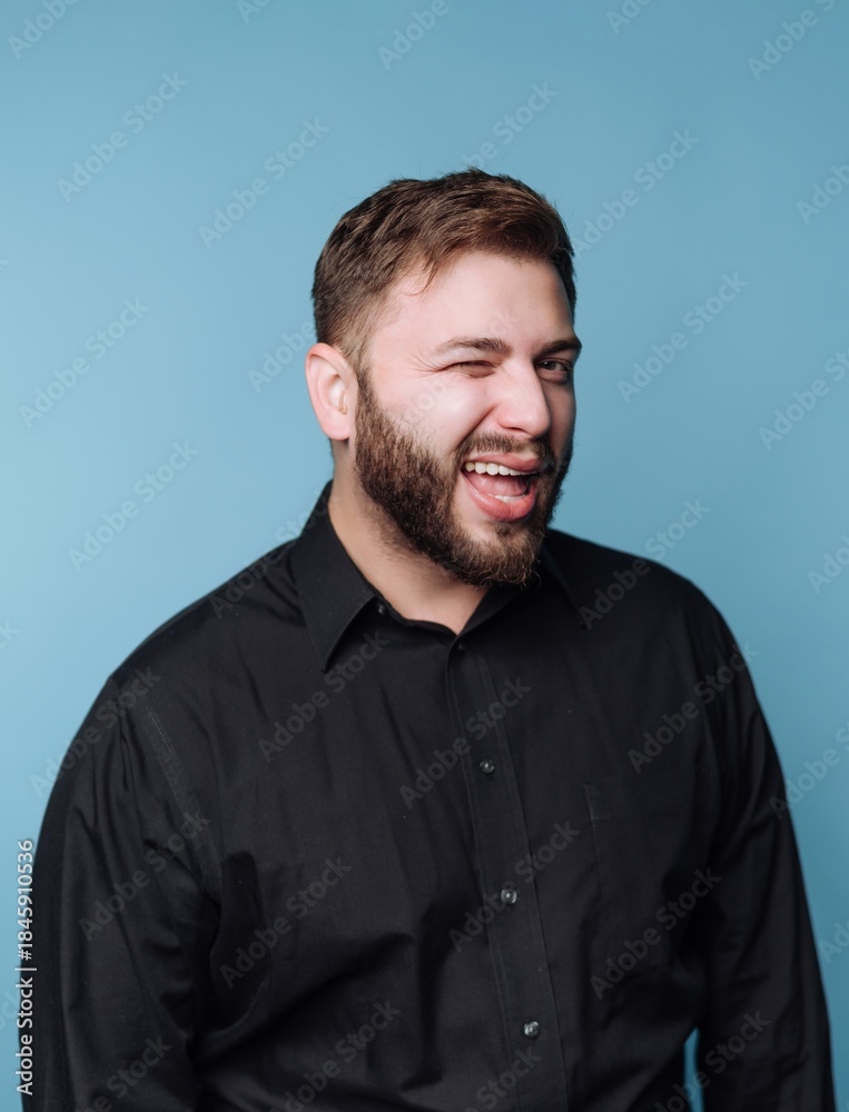 Obraz premium Man smiles and winks against a blue backdrop while wearing a black shirt and showing a playful expression