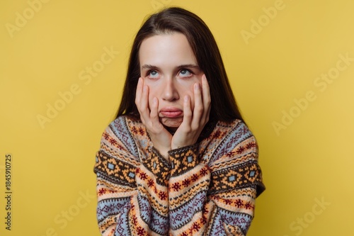 Young woman shows boredom while sitting against a yellow wall with her hands on her face