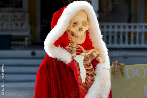 A human skeleton on a stand wearing a bright red soft faux fur cape and thick white hood. A body of bones wears the funny Santa Claus suit. The head has large eye socket holes and teeth dentures 