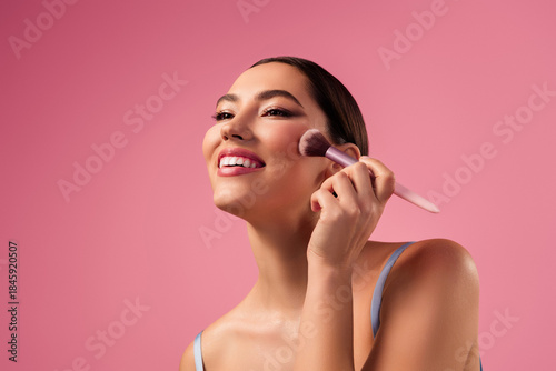 Gorgeous Young woman applying makeup with a brush, smiling gracefully against a pink background.
