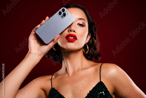Stylish young woman in a chic green sequin dress holding a smartphone set against a bold red backdrop