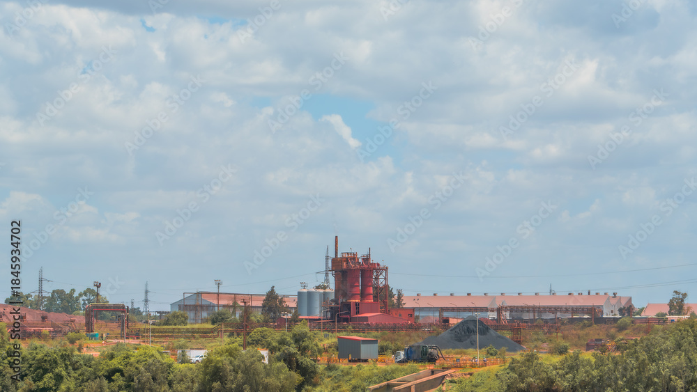 Obraz premium Industrial landscape featuring a red furnace and factory buildings, surrounded by green trees under a bright, cloudy sky