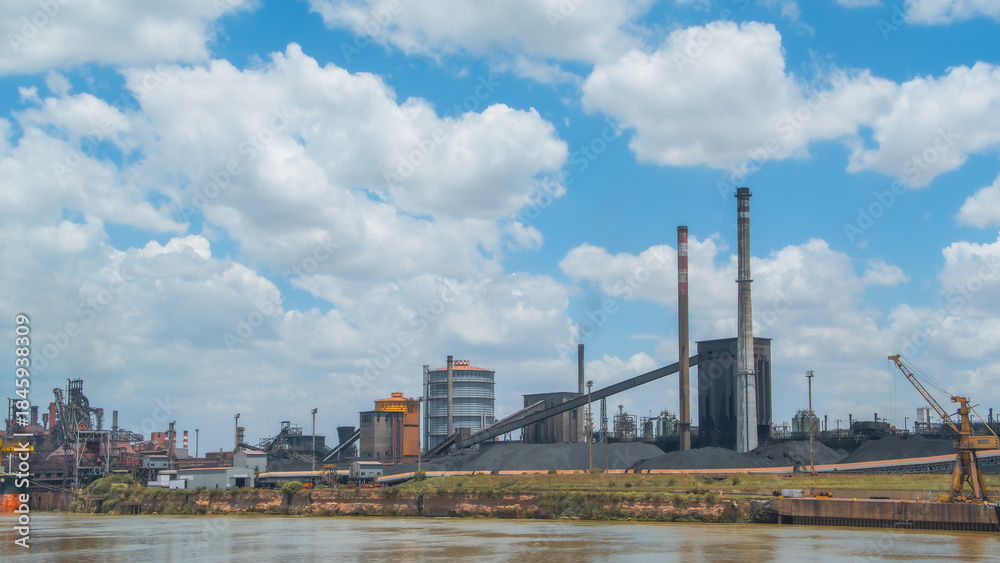 Obraz premium Industrial steel mill or factory complex with tall smokestacks, large conveyor belts, and piles of coal on a riverside under a vast cloudy blue sky
