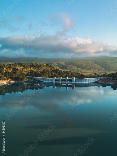 A serene aerial view of a reservoir dam in South Spain, surrounded by lush hills and calm water reflecting the sky, ideal for nature, travel, and infrastructure concepts.