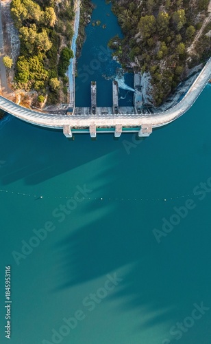 Drone photo of a dam and turquoise reservoir surrounded by forest in South Spain, ideal for illustrating water management, infrastructure, and environmental conservation topics.