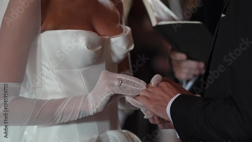 Bride Sliding Ring Onto Groom Finger During Vows, Gloved Hands And Satin Dress Visible While Officiant Holds Book Nearby Creating Solemn,