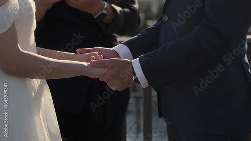 CloseUp Bride Groom Hands Exchanging Rings, Diverse Couple At Waterfront Ceremony With Officiant, Soft Backlight, Navy Suit And White