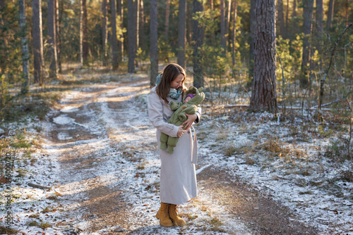 A girl with a child in a sunny winter forest. A mother holds her daughter in a park. The woman smiles at her newborn. The baby is wearing a winter jacket.