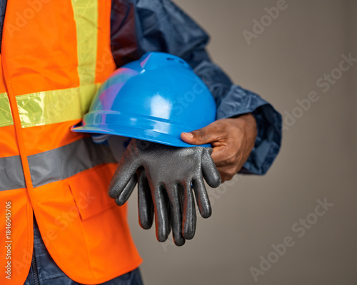 A close-up shows a worker wearing an orange reflective safety vest holding a blue hard hat and a pair of protective gloves against a neutral studio background.