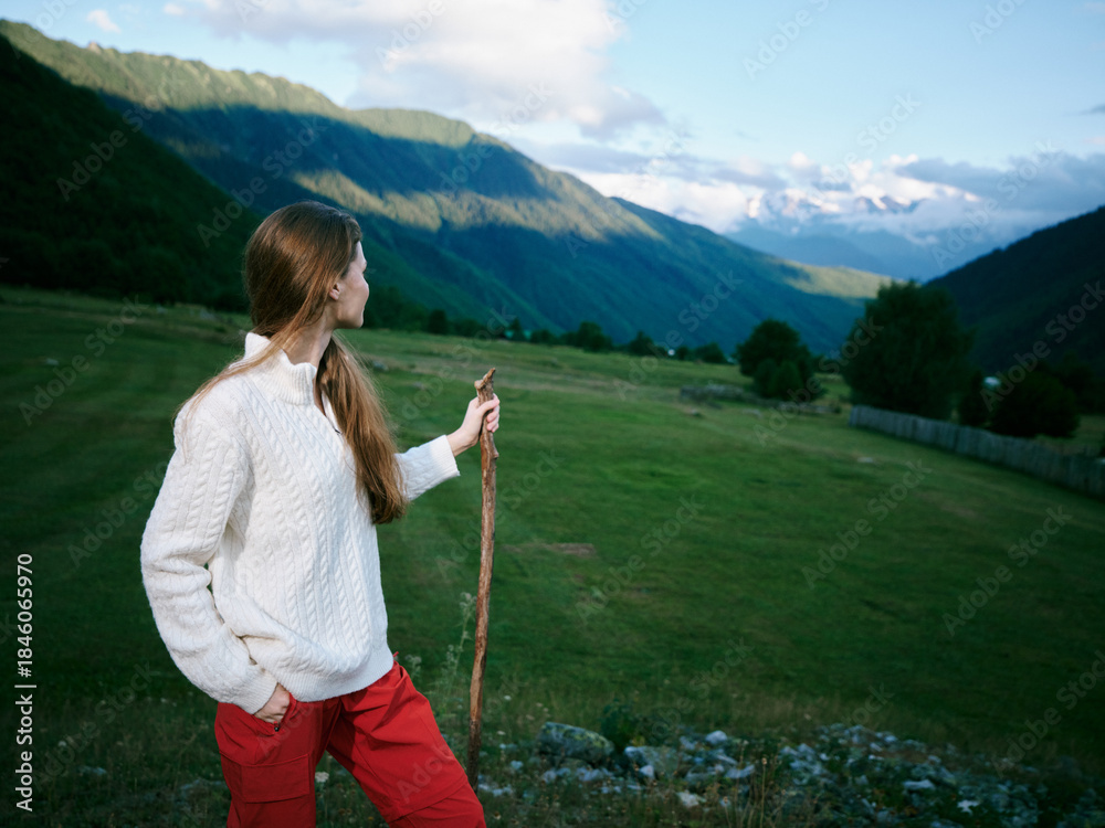 Fototapeta premium Woman in a white sweater stands with a walking stick in a lush valley, gazing toward distant mountains, an outdoor scene of nature, exploration, and peaceful countryside.