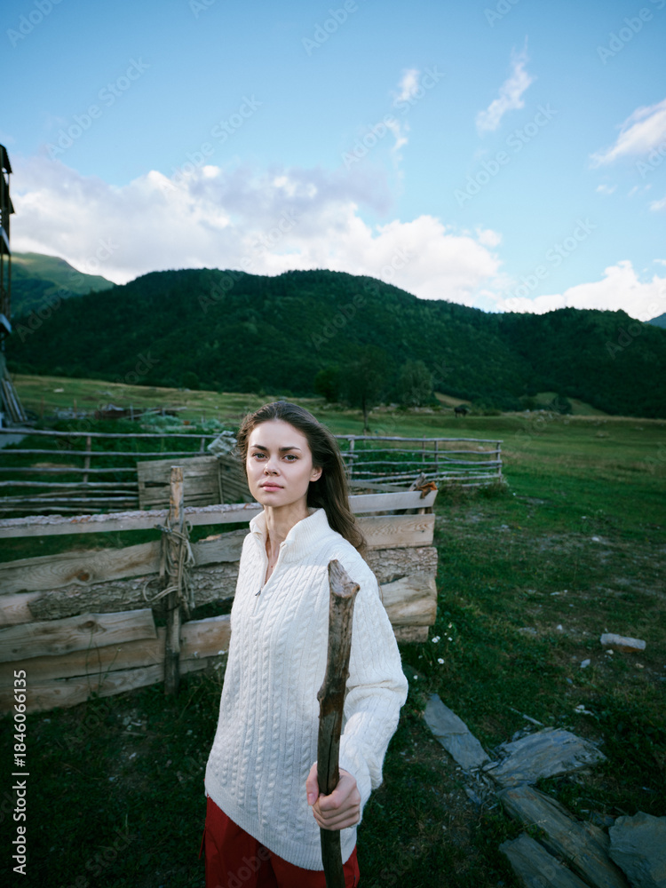 Fototapeta premium Woman in a white knitted sweater stands beside a wooden fence in a wide countryside field, carrying a wooden stick, with green hills and blue sky behind