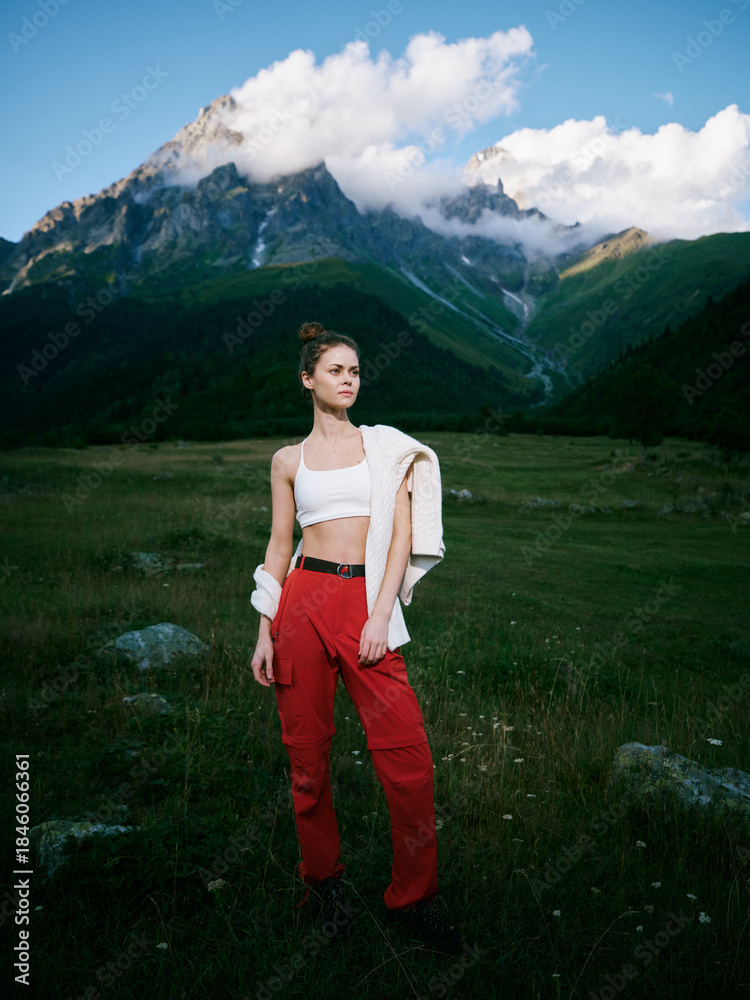 Fototapeta premium Mountain landscape with a stylish woman standing in a green meadow, wearing a white crop top and red pants, jacket draped over shoulder, crisp sky and distant peaks.