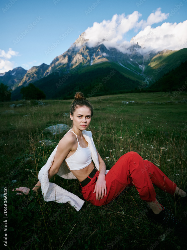 Fototapeta premium Woman sitting on a grassy meadow with mountains in the background, wearing a white top and red pants, enjoying outdoor nature, calm scenery, and fresh air.