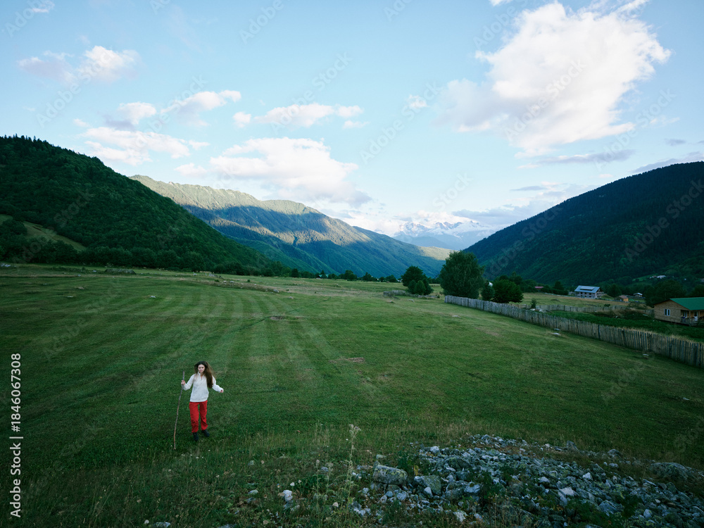 Fototapeta premium Wide meadow between hills with a girl walking across green grass, distant mountains under a bright sky and clouds, capturing freedom, nature, and outdoor exploration