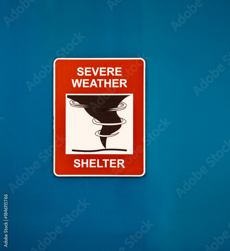 A red sign for a Severe Weather Shelter against a blue wall with a graphic of a tornado or hurricane