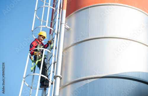 Safety-equipped worker climbing an enclosed ladder on a large industrial structure, representing regulated access and fall prevention systems.