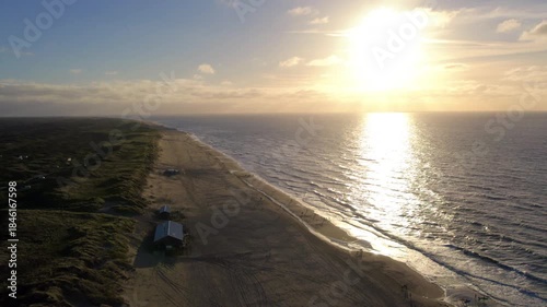 Aerial sunset over sandy coastline, cinematic drone sweep captures shimmering sea reflection and sun near horizon, gentle waves