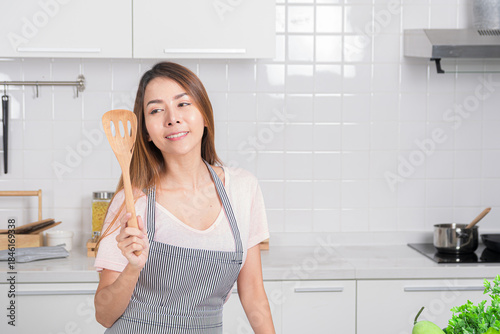 Beautiful Asian young woman joyfully poses in her white kitchen, holding cooking tool is wood spatula representing love for homemade meal.