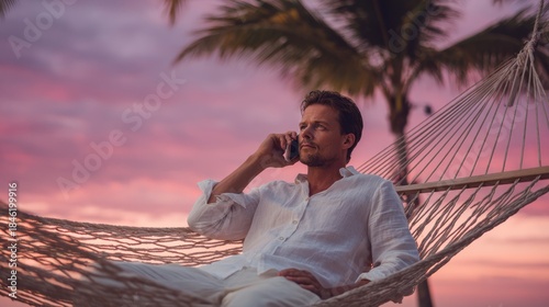 Man talking on a mobile phone while relaxing in a hammock at a tropical beach during a vibrant pink and purple sunset.