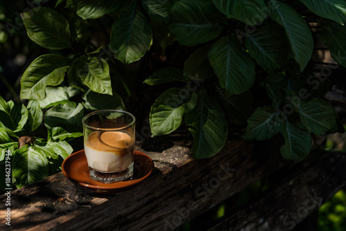 a small shot glass of chilled milk filled with hot espresso shot coffee floated over white creamy chilled milk put on wooden table in wild garden with sun shine and shade of green trees