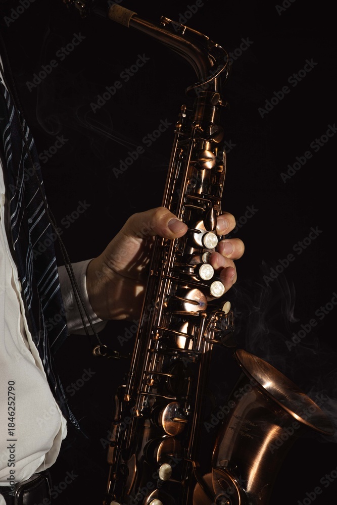 Obraz premium Close-up of a jazz musician’s hand holding a vintage brass saxophone against a dark background. Dramatic studio lighting highlights the instrument’s metal texture, keys, and curves. Moody atmosphere c