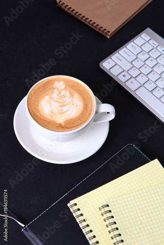 A cup of coffee on a black background. A notebook for writing and a computer keyboard on the table. The concept of a business breakfast or coffee break. Food and drinks.