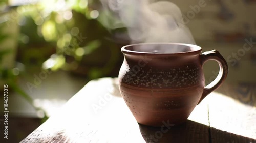 Steaming clay mug sits in sunlight with plants behind, on wood