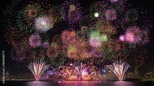 Colorful fireworks exploding over a vibrant city skyline at night, with reflections on the water surface.
