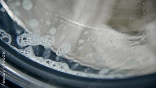 A close-up of a domestic washing machine window. White linens are washed, spinning and tumbling in the washing machine drum. Foam and bubbles run down the glass door viewing window.