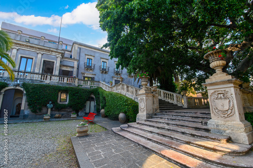 CATANIA, ITALY. The lush courtyard of Villa Cerami, a historic 18th-century palace currently housing the Faculty of Law, featuring a large palm tree, stone statues, and a grand stairc