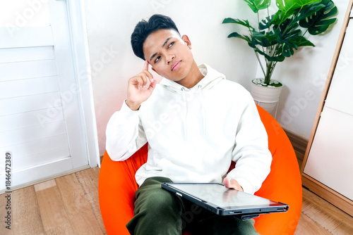 Young Asian Man Thinking While Using Laptop In Modern Living Room