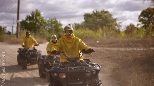 Yellow jacketed riders kicking up dust and gravel on ATV track, lead rider accelerates toward camera, dynamic motion and flying debris, cloudy sky, team chasing