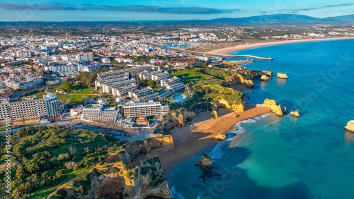 Aerial view of golden sands meet turquoise waters, cliffs rise dramatically, and buildings nestle along the coastline, Lagos, Faro, Algarve, Portugal.