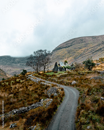 Aerial view of a winding road leading to a stone house nestled amidst the rugged, earthy tones of Killarney National Park under a muted sky, Killarney National Park, County Kerry, Ireland.