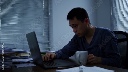An individual, deep in thought, hunched over a laptop at a desk. A coffee mug is present. Blinds are visible