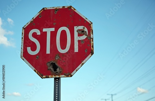 Damaged stop sign stands against blue sky. Rusty red roadsign is riddled with holes. Symbol of neglect and at city street warns drivers about safety.