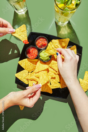 Female hands holding nachos corn chips and dipping them in salcha and guacamole sauces, glasses with lemonade are standing nearby. Mexican cuisine concept