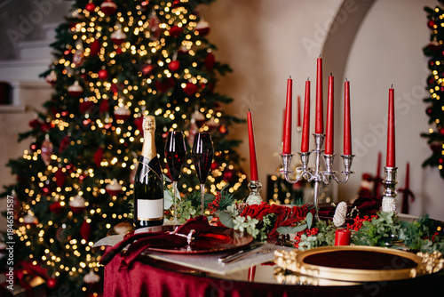 Christmas banquet table with candles, served with dishes, glasses and a bottle of champagne stands against the background of a Christmas tree with garlands