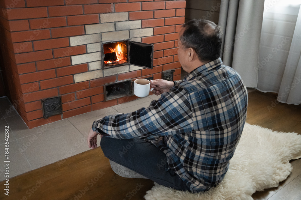 Obraz premium Man relaxing near burning fireplace while holding hot drink in white cup. Adult man sitting on floor in cozy home interior during cold winter season.