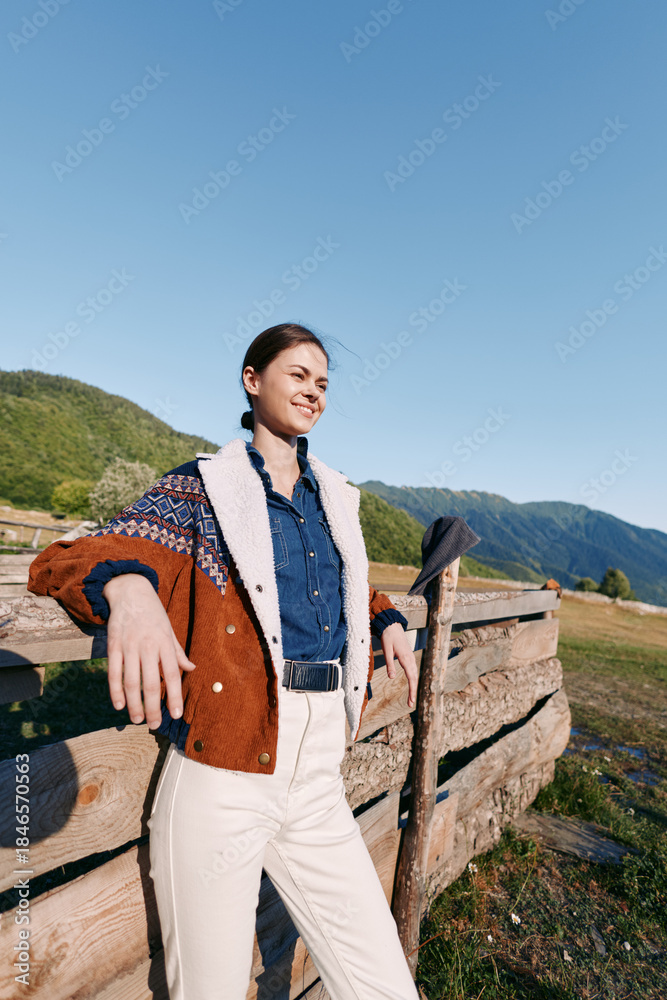 Fototapeta premium Woman in countryside leaning on wooden fence with mountains behind, outdoors portrait showing smile and relaxed casual cardigan style against blue sky and green meadow.