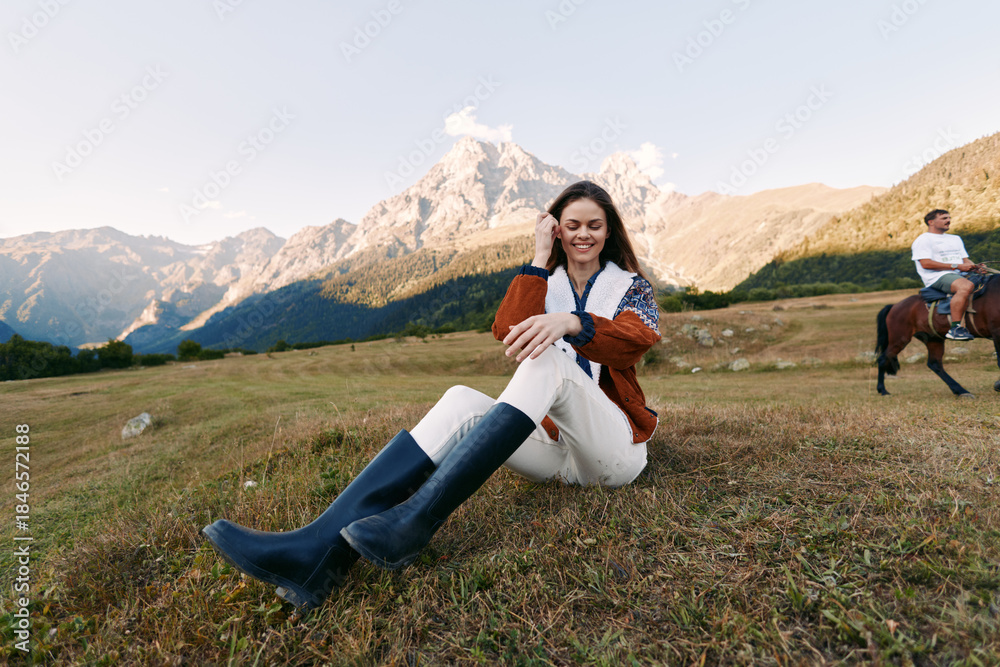 Fototapeta premium Woman in sweater and boots sits smiling on a grassy meadow with towering mountain peaks behind, outdoor portrait and leisure scene with horse and rider passing in the background.