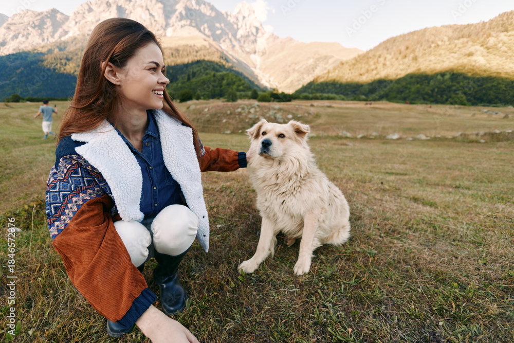 Fototapeta premium Woman petting dog in mountains meadow, smiling and crouching beside her pet in outdoor countryside scene with autumn light. Happy owner in warm jacket with furry companion.