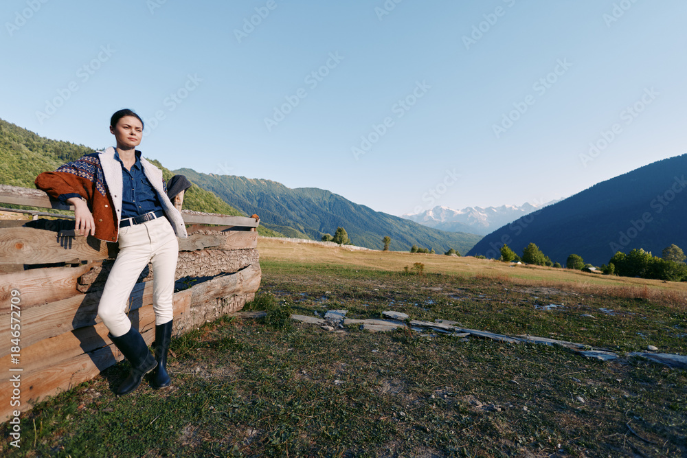 Fototapeta premium Woman in stylish jacket and boots leaning on wooden fence in mountainous meadow, enjoying countryside landscape and nature outdoors at golden hour, fashion portrait with distant peaks and open sky