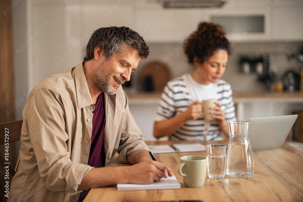 Fototapeta premium Couple At Kitchen Table Working Together: Man Writing Notes, Woman Drinking Coffee