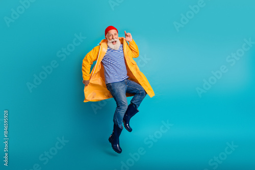 Foto Elderly man jumping in yellow raincoat with striped shirt beanie blue background