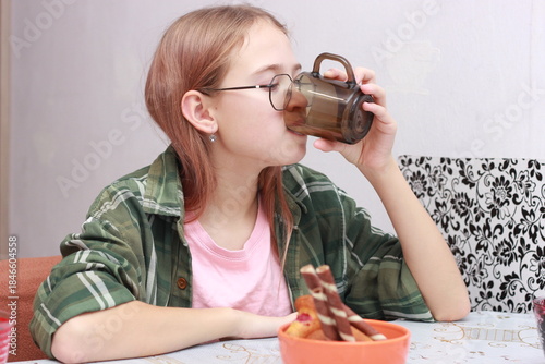 A teenage girl in a green shirt is drinking tea in the kitchen. Snack after school, breakfast.