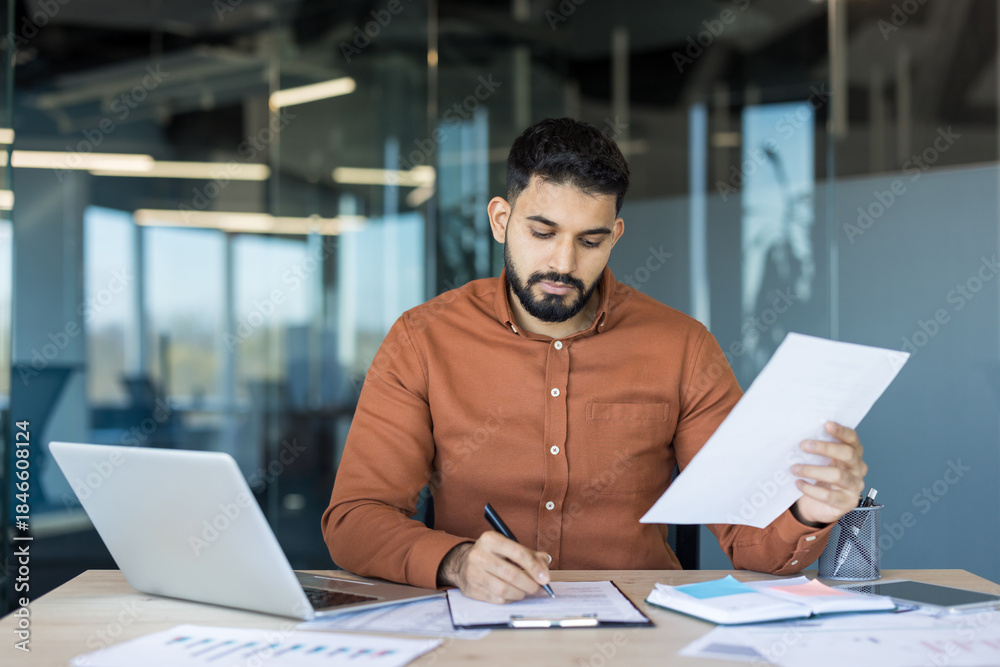 Fototapeta premium Businessman focused on reviewing and signing contract documents at a modern office desk with laptop and paperwork, conveying professional responsibility, negotiation and career-focused determination