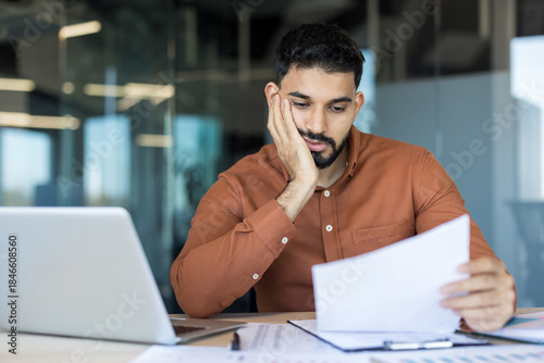 Stressed male employee experiencing frustration and overwhelm, holding document while feeling tired from heavy workload and endless paperwork in a modern office workplace