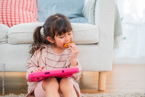 Young girl enjoying a cookie while sitting on the floor with a tablet in a cozy living room